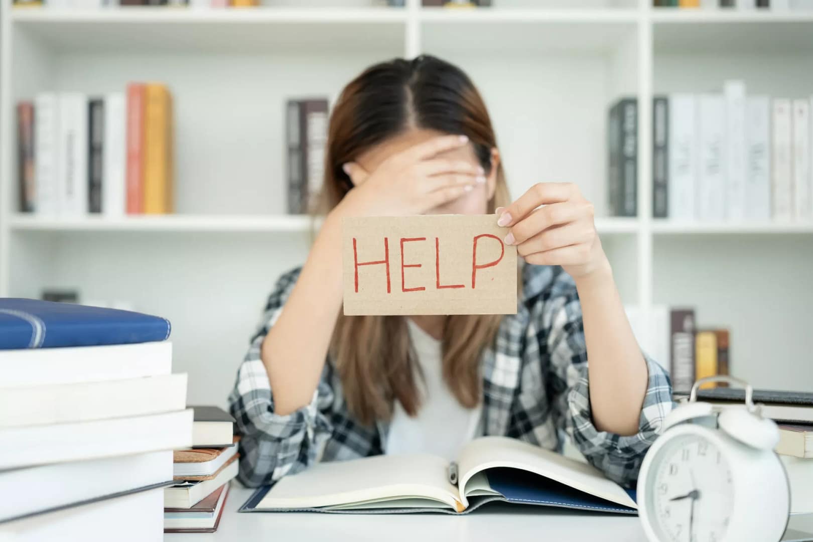 Une personne, peut-être un élève dyslexique en difficulté avec l’apprentissage de l’anglais, est assise à un bureau encombré, la tête dans une main, tenant un morceau de papier sur lequel est écrit HELP. Des livres et un réveil sont éparpillés sur le bureau tandis que des étagères remplies de livres se profilent à l’arrière-plan. Une personne, peut-être un élève dyslexique en difficulté avec l'apprentissage de l'anglais, est assise à un bureau encombré, la tête dans une main, tenant un morceau de papier sur lequel est écrit HELP. Des livres et un réveil sont éparpillés sur le bureau tandis que des étagères remplies de livres se profilent à l'arrière-plan.